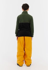 Fleece-Pullover mit grünem Oberteil und schwarzem Unterteil, kombiniert mit lockeren, gelben Cargo-Hosen, die einen hohen Kragen und eine strukturierte Stoffoberfläche aufweisen.