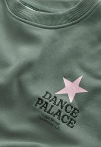 Sudadera verde con cuello de canalé, que presenta una estrella rosa y el texto "DANCE PALACE" en bordado negro. Textura suave.