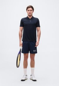 Νεαρός άνδρας που φοράει σκούρα μπλε αθλητικά ρούχα και λευκά αθλητικά παπούτσια, κρατώντας ρακέτα του τένις, στέκεται μπροστά σε ένα απλό ανοιχτόχρωμο φόντο.