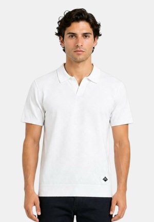 Uomo con capelli scuri che indossa una polo bianca semplice a maniche corte e pantaloni scuri, in piedi davanti a uno sfondo chiaro e uniforme.