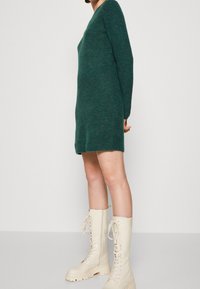 Abito maglione in un profondo teal con maniche lunghe e scollo a V. Abbinato a stivali da combattimento crema con lacci e suole robuste.
