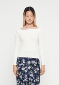 Weißes, geripptes Langarmoberteil mit quadratischem Ausschnitt, kombiniert mit einem blumengemusterten blauen Rock mit lila und grünen Akzenten.