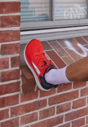 Roter Sportschuh mit strukturierter Oberfläche, schwarzen Akzenten und weißer Sohle, liegt auf einer Ziegeloberfläche in der Nähe eines Fensters. Weißer Socken sichtbar.