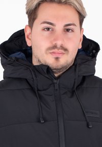 Czarna kurtka puchowa z kapturem, z zapięciem na suwak z przodu, regulowanymi sznurkami oraz subtelną naszywką logo na klatce piersiowej.