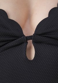 Μαύρο μαγιό με υφή, σε σχέδιο κυψελωτής δομής. Διαθέτει λαιμόκοψη σε διακοσμητική καμπύλη και λεπτομέρεια σε σχήμα κλειδώματος στο κέντρο, δεμένο στη βάση.