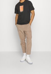 Svart T-shirt med grafisk tryck, beige kordflosbyxor och vita sneakers. Modellen står med händerna i fickorna och visar upp en avslappnad outfit.