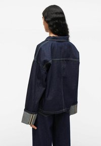 Vrouw met lang zwart haar draagt een donkerblauwe spijkerjasje en bijpassende broek, met de rug naar de camera gekeerd tegen een effen witte achtergrond.