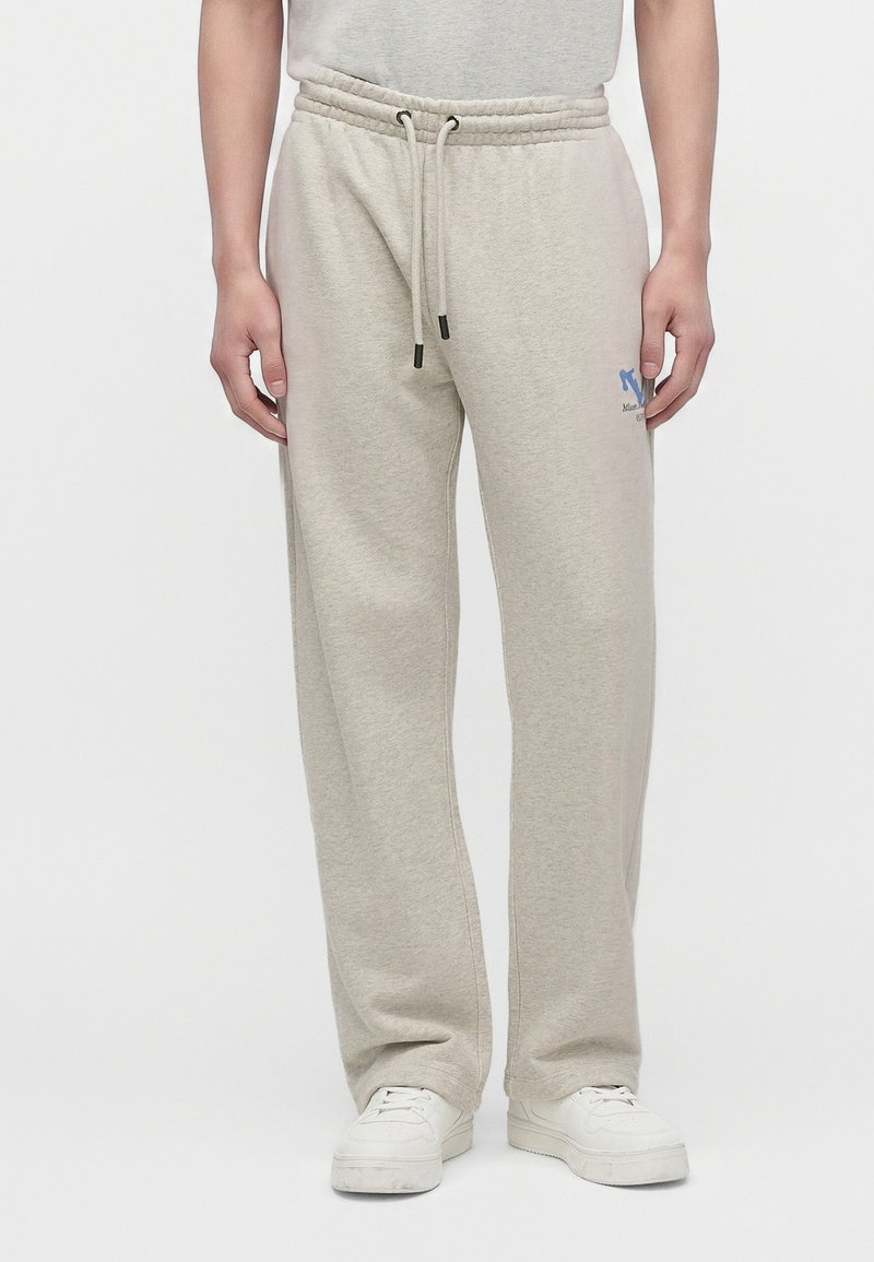 Homme portant un pantalon de jogging beige clair avec taille à cordon et des baskets blanches, debout devant un fond uni.