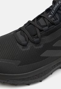Κοντινή λήψη μαύρου παπουτσιού Terrex για τρέξιμο σε μονοπάτι με επάνω μέρος από διχτυωτό υλικό, χοντρή υφή στην σόλα και σταυρωτά κορδόνια.