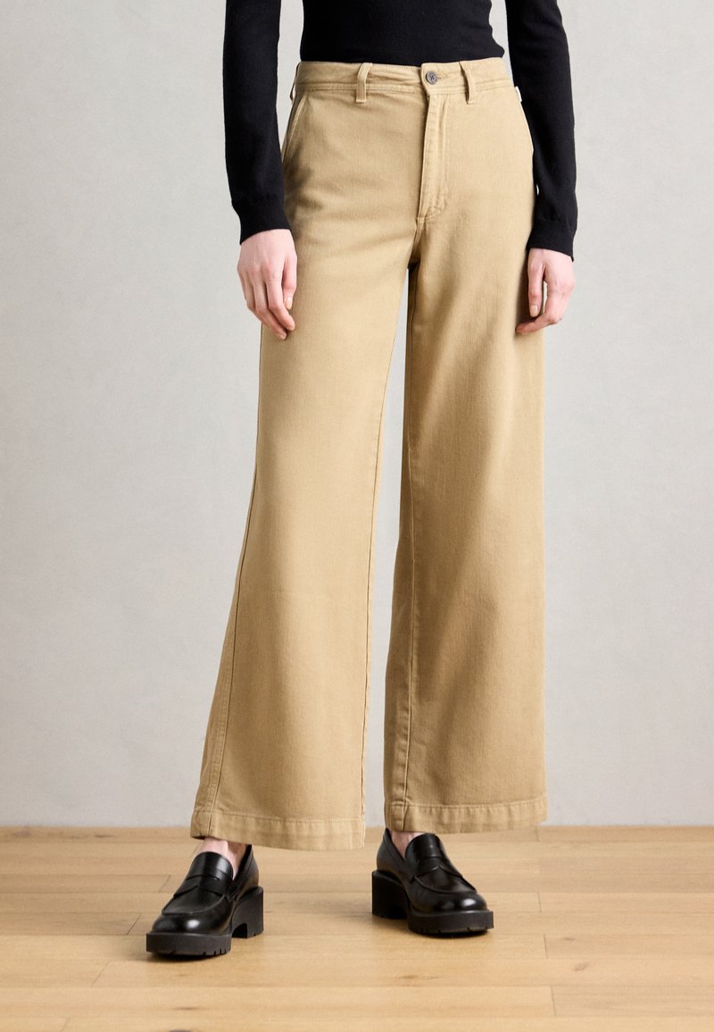 Beige vide benbukser laget av bomull, med høy midje, klassiske frontlommer og rett ben, parret med svarte loafers.