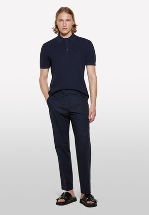 Polo shirt bleu marine à manches courtes avec un motif texturé, associé à un pantalon habillé bleu marine et des sandales noires avec un accent en nœud.