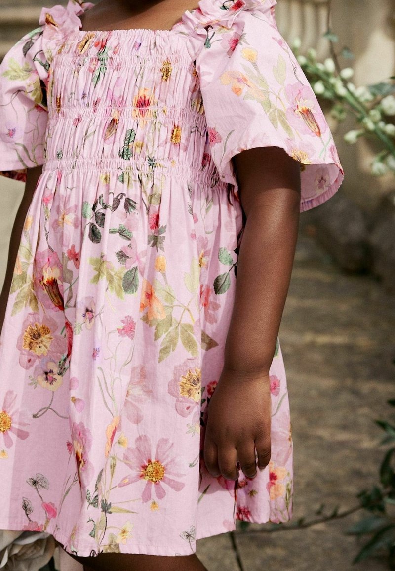 Enfant portant une robe rose fleurie avec des manches bouffantes, debout à l'extérieur avec de la verdure en arrière-plan, main détendue le long du corps.