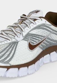 Λευκό και καφέ παπούτσι τρεξίματος Nike με υφή πλέγματος, καφέ λογότυπο swoosh, λευκά κορδόνια και τμηματική λευκή σόλα.