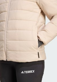 Nahaufnahme einer Person, die eine hellbeige Steppjacke mit Reißverschluss und schwarze TERREX Leggings trägt, mit den Händen in den Taschen der Jacke.