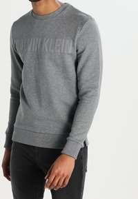 Sudadera gris con cuello redondo, puños y dobladillo acanalados, que presenta un destacado logo de Calvin Klein en un tono más oscuro en el pecho.