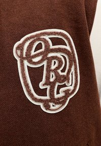 Brunt stoff med en strukturert hvit og brun brodert logo som viser sammenflettede bokstaver. Designet har en lagdelt, myk tekstur.