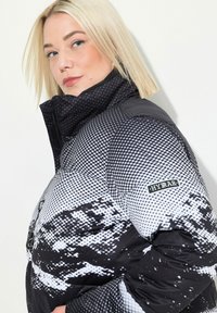 Chaqueta acolchada en blanco y negro con un patrón pixelado, cuello alto y parche con logo. El material es brillante, proporcionando una textura reluciente.