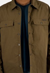 Giacca button-up marrone con due tasche sul petto e bottoni a pressione grigi, indossata sopra una maglietta a V blu scuro. Tessuto di texture liscia.