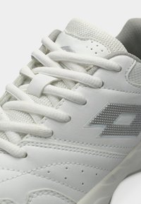 Λευκό αθλητικό παπούτσι με λείο επάνω μέρος από δέρμα, με λεπτομέρειες από δίχτυ, επίπεδα κορδόνια και ανάγλυφο λογότυπο στο πλάι. Τρύπες για καλύτερο αερισμό.