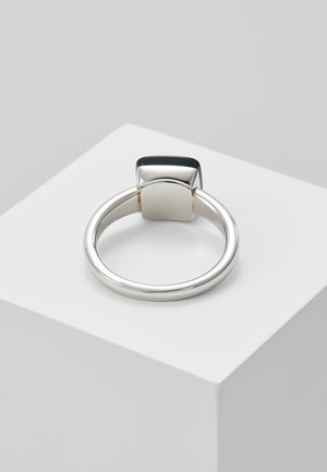 Zilveren ring met een rechthoekige zwarte edelsteen gezet in een gladde, gepolijste band die ligt op een witte geometrische ondergrond.