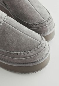 Grå ruskind slip-on sko med afrundet tå, detaljeret syning langs sømmen og en tekstureret gummisål for bedre greb.