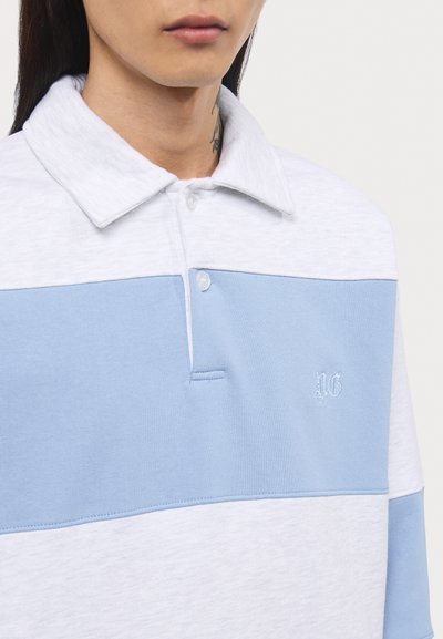 Polo-skjorte i lys grå og blå, laget av bomull. Den har krage med knapper, kontrasterende horisontale fargeblokker, og brodert logo på brystet.