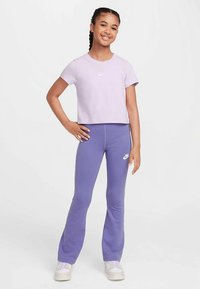 Violetinė trumpa marškinėliai su mažu baltu logotipu, derinami su violetinėmis plačiomis tamprėmis su baltu logotipu, dėvimi su baltais sportbačiais.