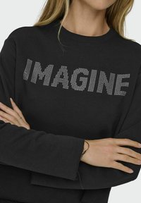 Hanorac negru cu cuvântul "IMAGINE" din ținte argintii. Hanoracul are un design lejer și mâneci lungi cu un detaliu de tiv brut.