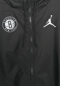 Sort vindjakke med en glansfuld overflade, med lynlås foran, justerbar hætte og hvide logoer: Brooklyn Nets og Jumpman-emblem.