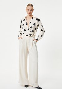 Blusa avvolgente bianca con grandi pois neri, abbinata a pantaloni crema a vita alta. La blusa presenta un profondo scollo a V e maniche lunghe.