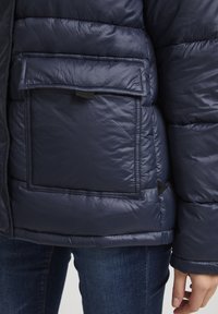 Tamsiai mėlynas pūkinis švarkas su didele kvadratine kišenę, siuvinėtu dizainu ir lygiu paviršiumi. Yra mažas etiketė apatinėje siūlėje.
