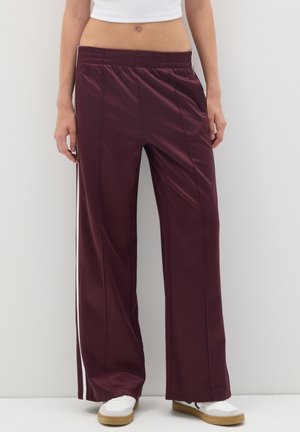 Person mit weinroten, weit geschnittenen Jogginghosen mit weißen Seitenstreifen, einem weißen Cropped-Top und weißen Sneakers mit Gummisohlen.