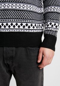 Czarnobiały sweter na drutach z geometrycznymi i wzorzystymi motywami, z prążkowanymi mankietami i dolnym brzegiem, noszony na ciemnych dżinsach.