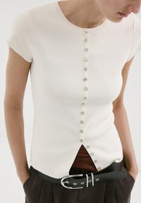 Femme portant un haut blanc ajusté à manches courtes avec une rangée verticale de boutons, associé à un pantalon foncé et une ceinture noire avec une boucle argentée.