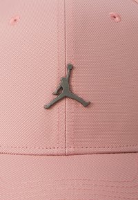 Lyserød kasket lavet af struktureret stof med et sølv Jumpman-logo på fronten. Enkel design med en buet skygge og stygningsdetaljer.