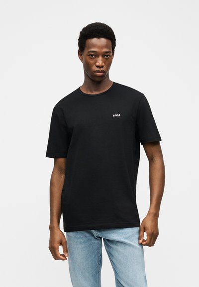 Jeune homme portant un t-shirt noir avec le logo "BOSS" sur la poitrine et un jean bleu clair, debout devant un fond clair uni.