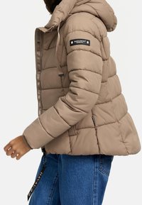 Lichtbruine gewatteerde jas met capuchon, doorgestikte textuur, ritssluiting en een gebrande patch op de mouw. Gecombineerd met een blauwe spijkerbroek.