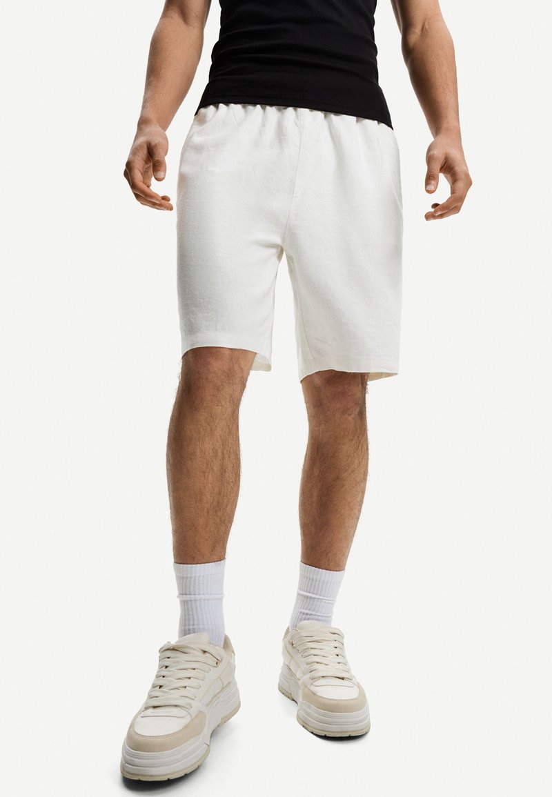 Homme portant un short blanc mi-genoux, des chaussettes blanches, des baskets beige et blanches, et un haut noir sans manches sur un fond uni.