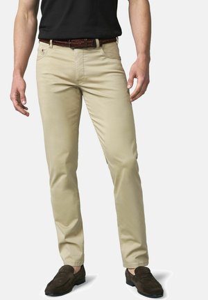 Mann trägt beige Slim-Fit-Hose, schwarzen Gürtel, schwarzes Hemd und dunkelbraune Wildleder-Loafer, steht vor einem einfarbigen hellen Hintergrund.