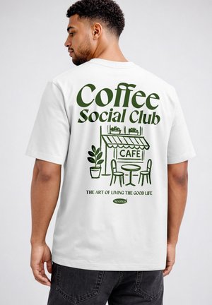 Man wearing white T-shirt with green text "Coffee Social Club" and cafe illustration with table, chairs, and plants on the back.
