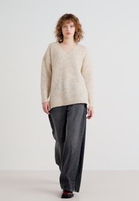 Beige bordázott kötött pulóver V-nyakkal, széles szárú fekete farmernadrággal és fekete cipővel párosítva, laza szabással és ejtett vállakkal.