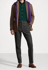 Hnědý herringbone blazer, tmavošedé kalhoty, zelená košile s knoflíky a fialový svetr přehozený přes ramena. Hnědé kožené mokasíny.