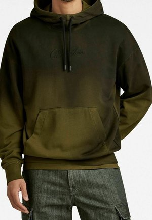 Homme portant un sweat à capuche olive de G-Star Raw avec poche avant et pantalon cargo en denim gris, posant devant un fond blanc.