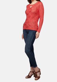 Czerwony sweter dziany z teksturowanym wzorem, długimi rękawami i detalem z klamrą przy dekolcie, zestawiony z ciemnymi dżinsami i brązowymi sandałami na obcasie.