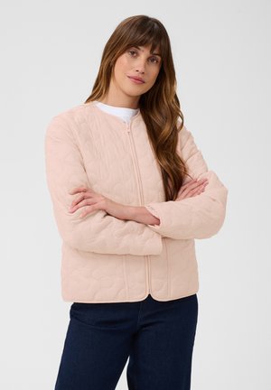 Femme aux longs cheveux bruns portant une veste matelassée rose clair avec une fermeture éclair sur le devant et un pantalon bleu foncé, debout les bras croisés.