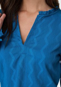 Blouse bleue texturée avec un motif en zigzag, décolleté en V et des accents volantés au col et aux poignets. Le matériau a une finition froissée.