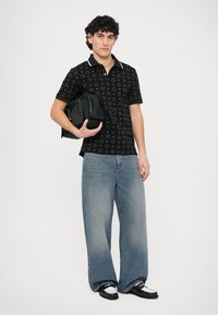 Polo preta com padrão estampado, gola branca. Jeans azul-claro de perna larga, bolsa preta na mão, sapatos pretos com detalhes brancos.