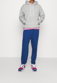 Felpa grigia con cappuccio, sottogiacca rosa, pantaloni della tuta blu e sneakers multicolori. Design sportivo casual, texture morbida e dettagli con cordini.