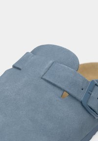 Μπλε σουέντ παπούτσι slip-on με ρυθμιζόμενο λουράκι, ανοιχτό σχέδιο στα δάχτυλα και υφή στην επιφάνεια. Διαθέτει εσωτερική επένδυση σε καφέ απόχρωση.