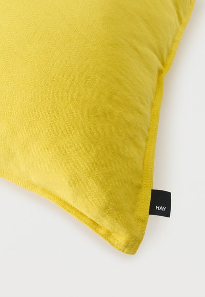 Coussin rectangulaire en tissu jaune vif avec texture visible et une petite étiquette noire portant la mention « HAY » sur une surface blanche.